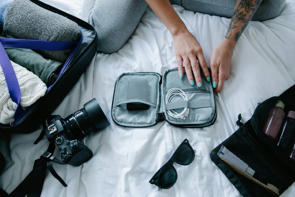 "A traveler packing a suitcase on a bed, organizing charging cables in a grey tech pouch alongside a DSLR camera, sunglasses, and a toiletry bag. electronics, camera, and sunglasses in a suitcase on a bed.
