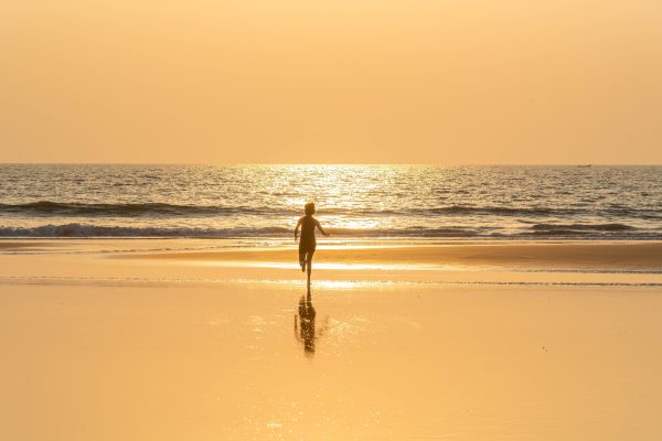 A child runs along a Goan beach during a golden sunset, casting a reflection on the wet sand.